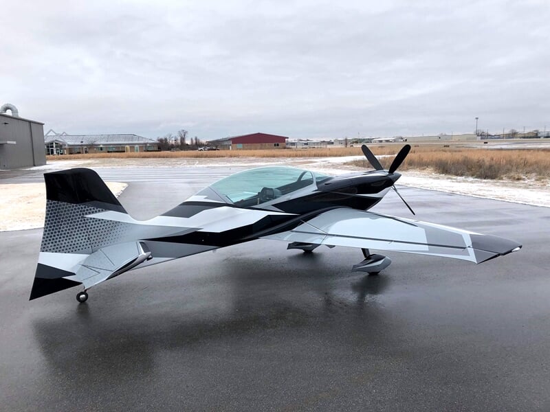 Petit avion de voltige noir et blanc stationné sur une piste mouillée sous un ciel gris.