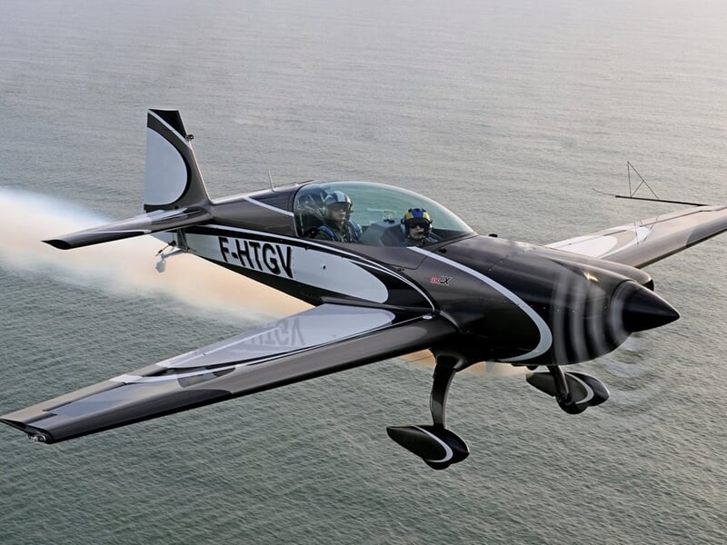 Avion de voltige noir et blanc en vol au-dessus de la mer avec hélice en rotation.