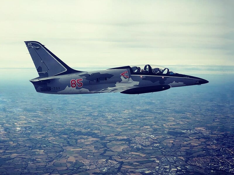 Avion de chasse Albatros L-39 gris avec le numéro 85 en vol au-dessus d'un paysage rural vu depuis le ciel, ciel clair et nuageux.