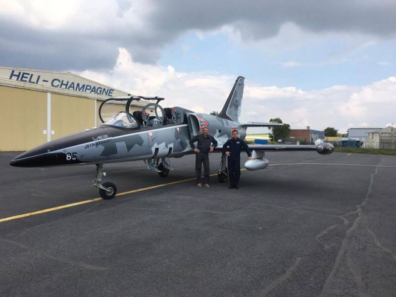 Deux personnes en combinaison posent debout devant un avion de chasse camouflé sur une piste d'aéroport.