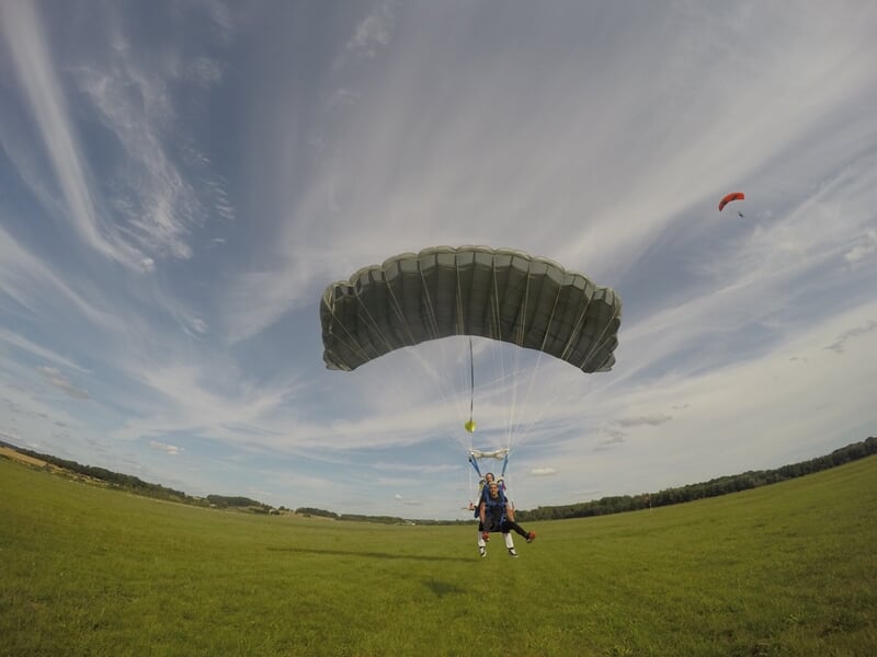 Un parachutiste atterrit en douceur sur un champ vert sous un ciel partiellement nuageux avec un autre parachute au loin.
