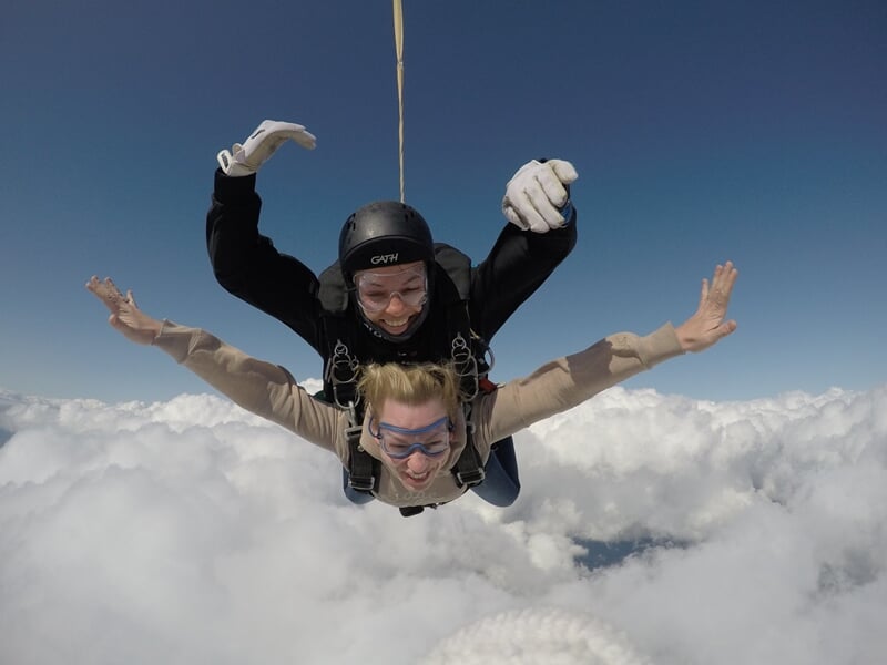 Deux parachutistes en tandem volent au-dessus des nuages blancs avec un ciel bleu en arrière-plan.