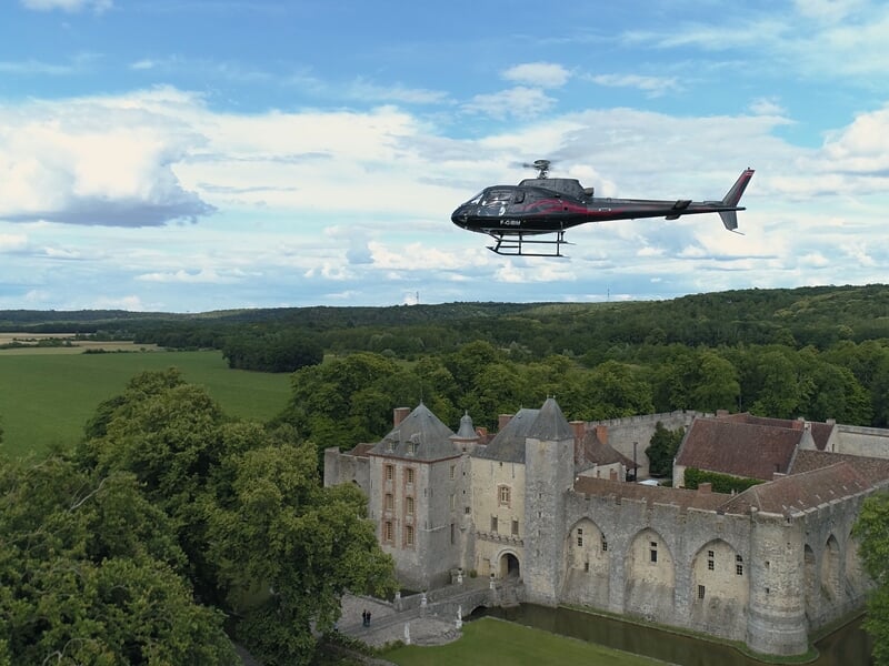 Hélicoptère noir volant au-dessus d'un château ancien entouré de forêt et de champs verts.
