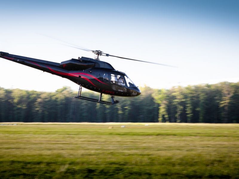 Hélicoptère noir avec des motifs rouges volant au-dessus d'un champ vert devant une forêt dense.