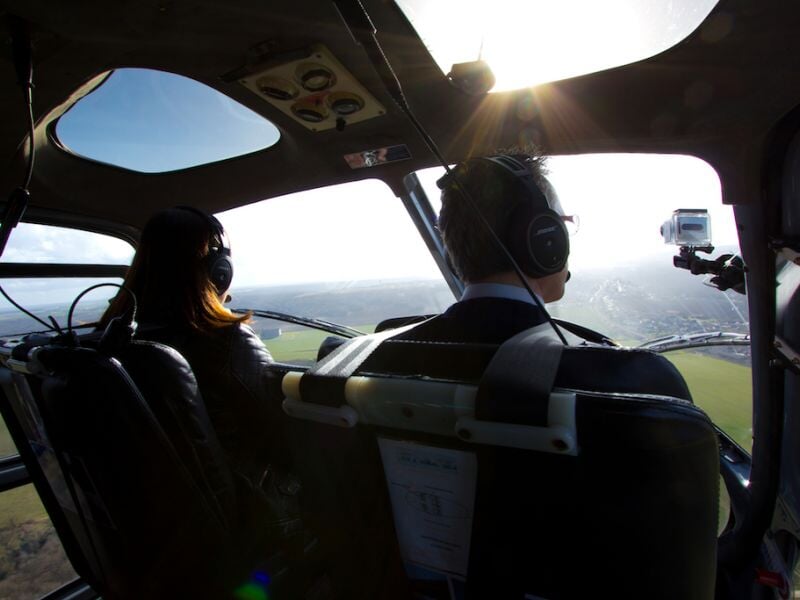 Intérieur d'un hélicoptère avec deux passagers portant un casque audio, vue sur le paysage en vol.
