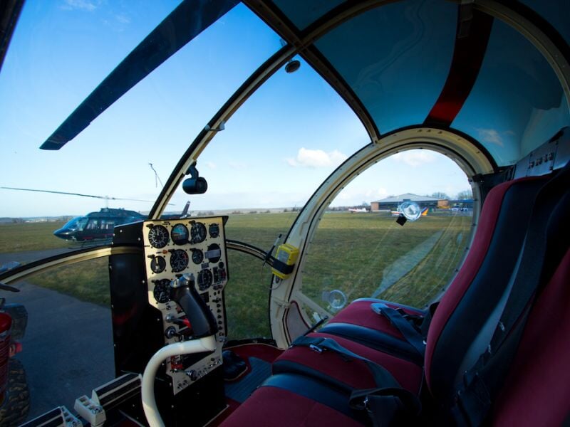 Cockpit d'hélicoptère avec tableau de bord et sièges rouges, vue sur un autre hélicoptère au sol.