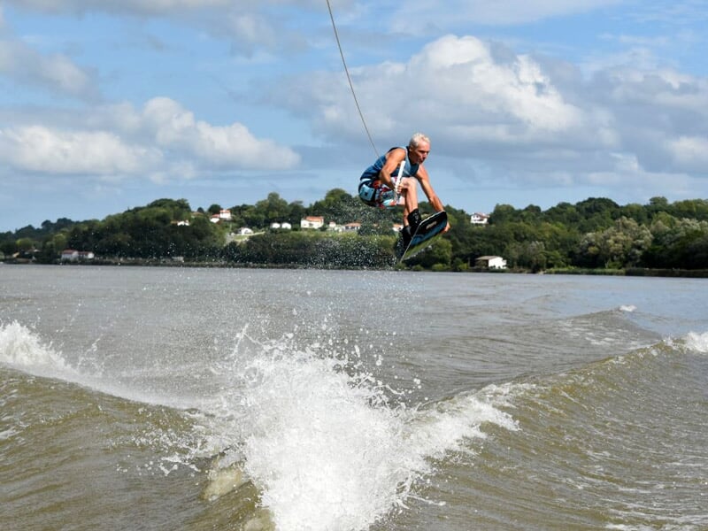 Personne en wakeboard en plein saut au-dessus de l'eau, avec un paysage boisé en arrière-plan.