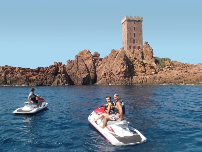 Groupe de personnes sur des jet skis rouges et blancs, près d'une tour en pierre sur des rochers rouges, mer calme et ciel bleu.