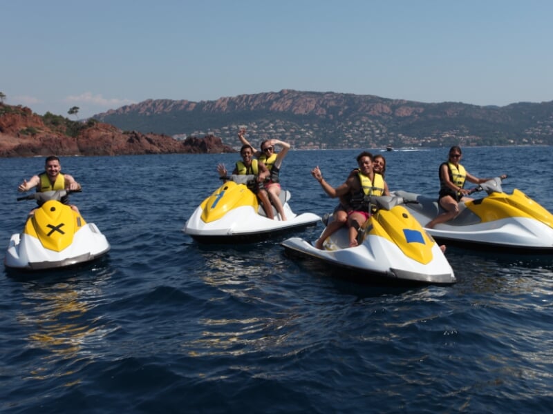 Personnes en gilet de sauvetage jaune faisant du jet-ski sur une mer calme, devant un paysage montagneux avec végétation méditerranéenne. Elles semblent s'amuser sous un ciel dégagé, certaines levant 