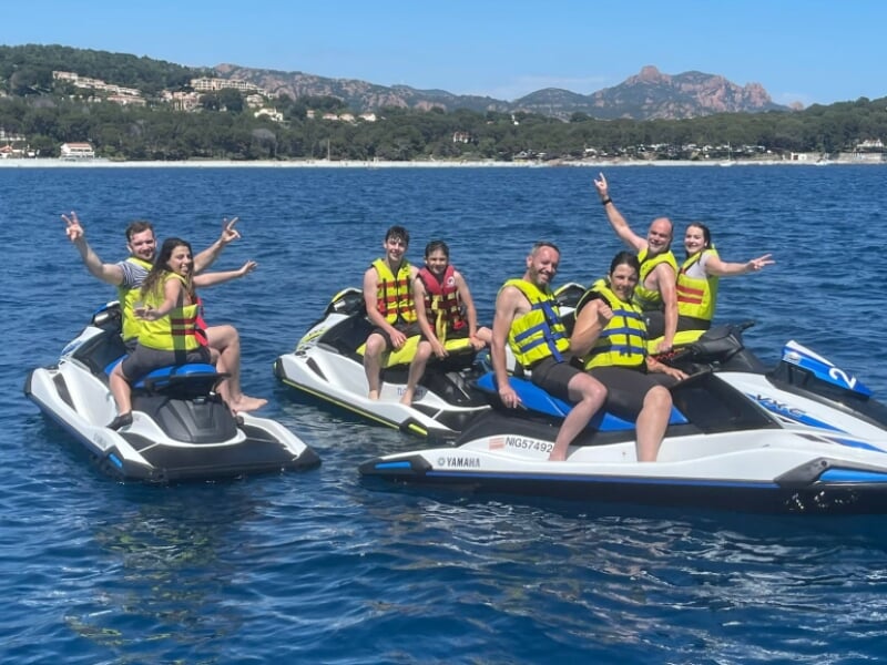 Groupe de personnes souriantes en gilet de sauvetage sur des jet-skis Yamaha, flottant sur une eau bleue transparente avec une plage et des collines boisées en arrière-plan. L'ambiance est conviviale 