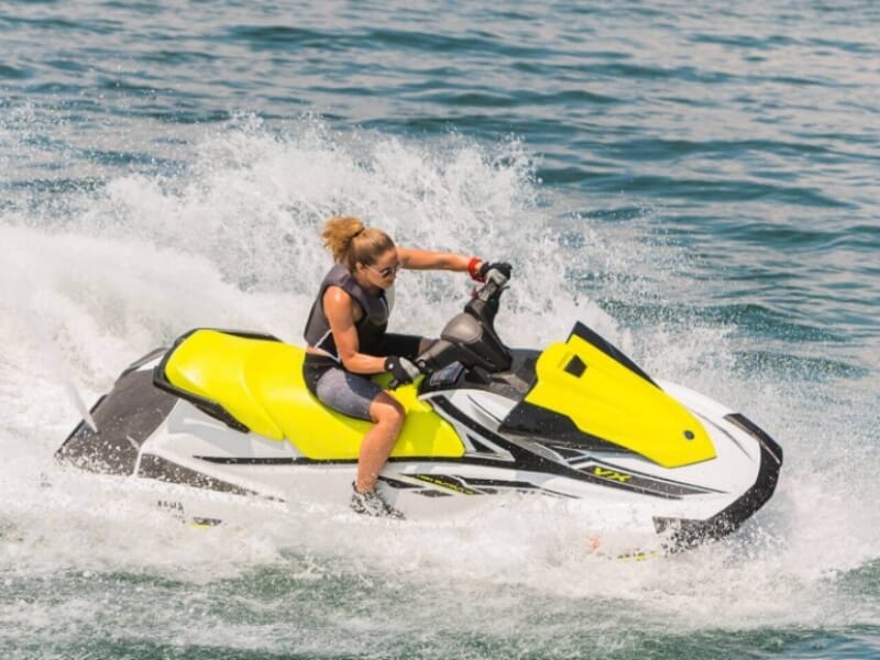 Femme en combinaison noire et jaune pilotant un jet-ski à grande vitesse, créant des éclaboussures d'eau autour d'elle. Concentrée, elle profite de l'activité sous un ciel ensoleillé.