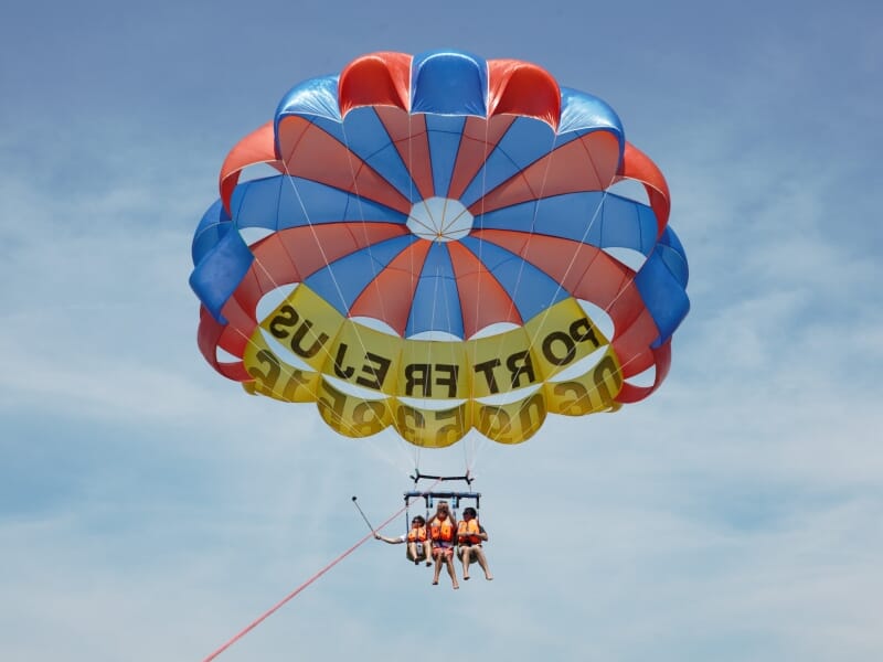 Trois personnes en parapente ascensionnel sur une mer calme, attachées à un parachute multicolore rouge, bleu et jaune, avec des gilets de sauvetage orange. Ciel bleu dégagé en arrière-plan, parfait p