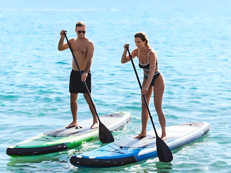 Un homme torse nu et une femme en maillot de bain, debout sur des planches de paddle sur une mer calme et bleue, pagayant côte à côte sous un ciel dégagé, pratiquant le stand-up paddle.