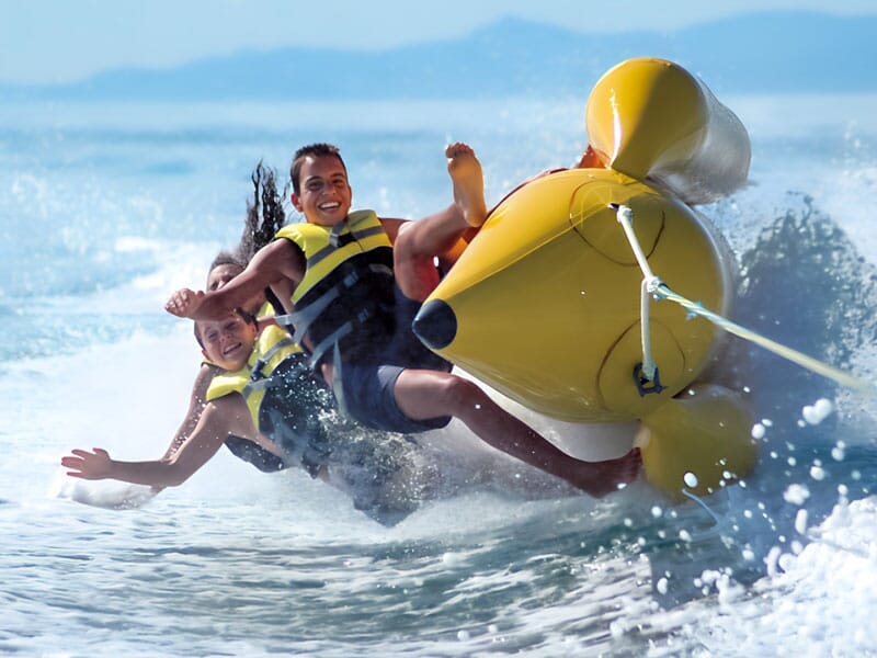 Deux personnes portant des gilets de sauvetage tombant d'une bouée gonflable jaune en pleine vitesse sur l'eau, avec des expressions de joie et d'excitation, éclaboussures d'eau autour d'eux.