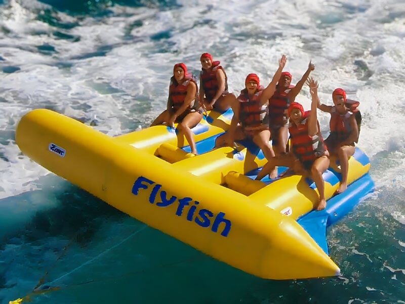 Groupe de personnes portant des gilets de sauvetage et des casques rouges, assises sur une bouée gonflable jaune Flyfish en mouvement rapide sur l'eau, levant les bras en l'air, souriants et profitant