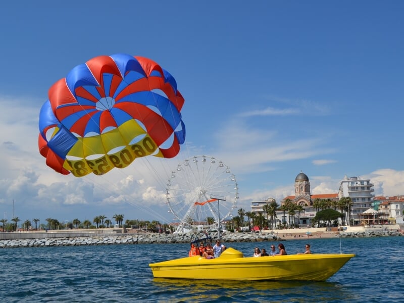 Plusieurs personnes sur un bateau jaune tirant un parachute ascensionnel bleu et rouge au large de la côte, une grande roue en fond