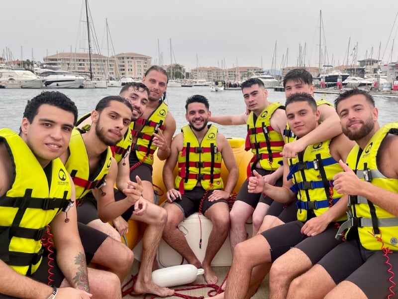 Groupe de neuf hommes en gilets de sauvetage jaunes assis sur un bateau amarré dans un port, entouré de yachts et de bâtiments, souriant et levant les pouces.