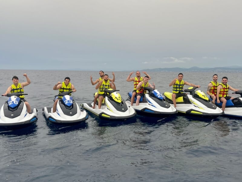 Six hommes en gilets de sauvetage jaunes chevauchent des jetskis blancs numérotés sur une mer calme, bras levés en signe de victoire, avec des montagnes en arrière-plan.