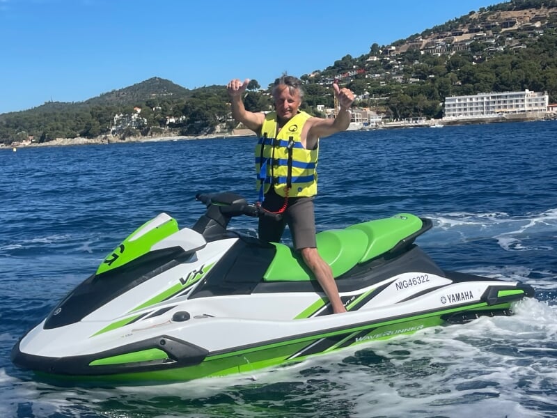 Homme en gilet de sauvetage jaune sur un jetski vert et blanc Yamaha Waverunner, levant les pouces, avec une côte boisée et des maisons en arrière-plan.