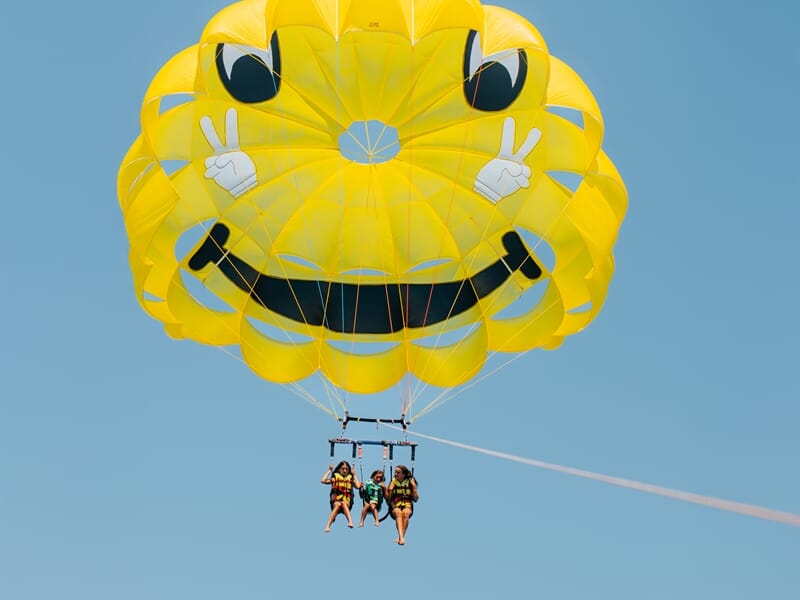 Trois personnes en parachute ascensionnel sous un parachute jaune souriant avec des mains faisant le signe de la paix.
