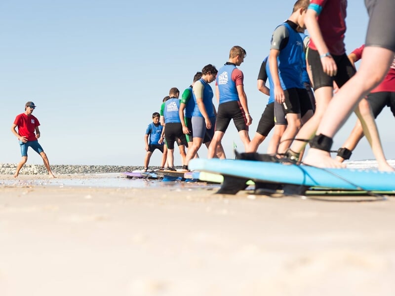 Groupe de jeunes surfeurs en combinaison bleue s'exerçant sur la plage avec un instructeur en rouge.