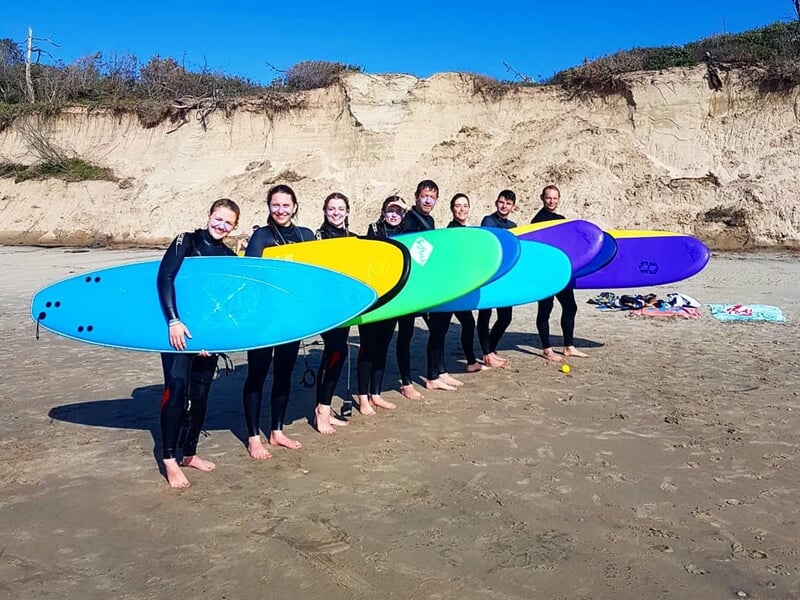Huit surfeurs souriants alignés sur la plage avec des planches colorées devant une falaise.
