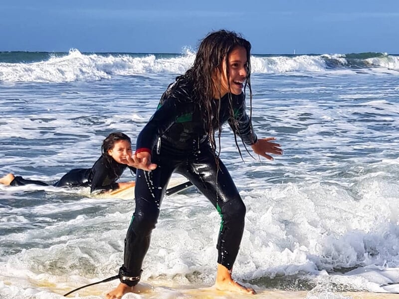 Jeune surfeuse souriante debout sur une planche dans les vagues avec une autre personne allongée dans l'eau.