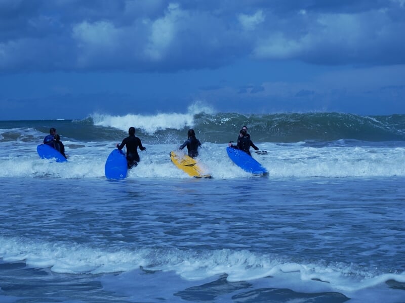 Trois surfeurs en combinaison noire pagayant sur leurs planches bleues et jaunes dans une mer agitée.