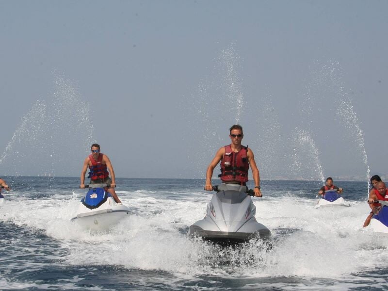 Quatre personnes conduisent des jetskis sur une mer calme sous un ciel clair, éclaboussant l'eau autour d'eux.