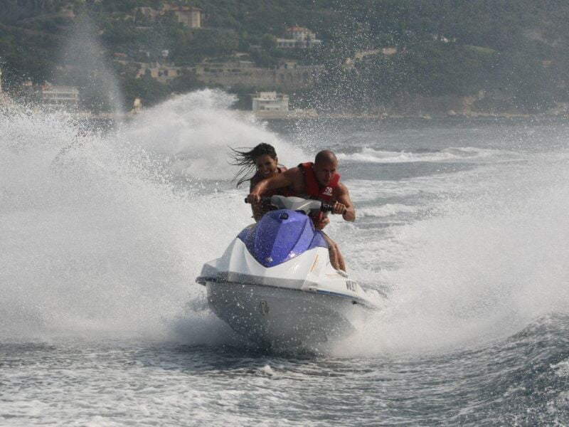 Un couple sur un jetski blanc et bleu fait une virée rapide sur l'eau, projetant une grande gerbe d'eau derrière eux.