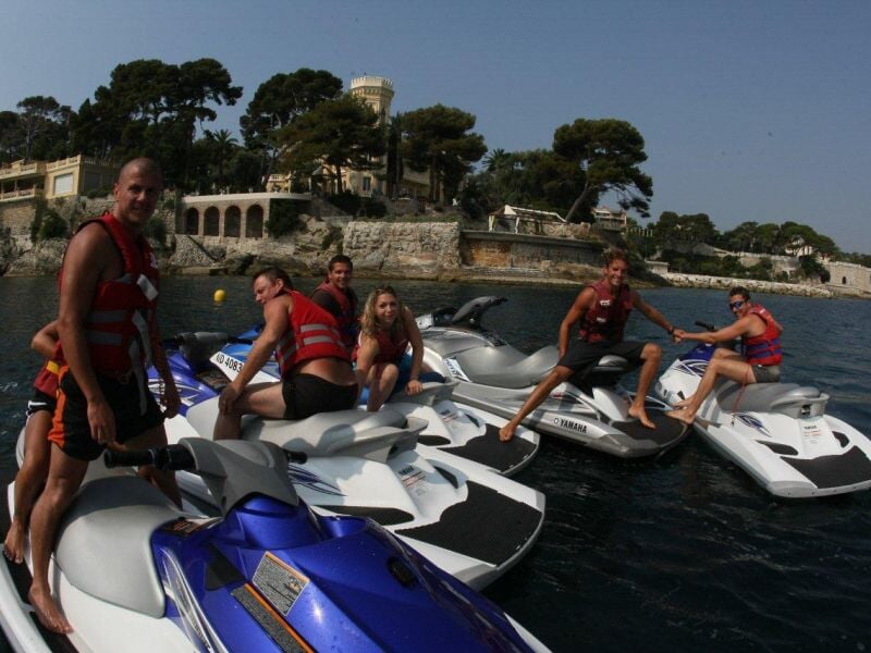 Groupe de six personnes en gilets de sauvetage posant sur plusieurs jetskis amarrés près d'une côte rocheuse avec des bâtiments.