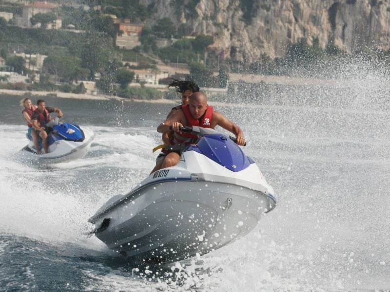 Deux personnes sur un jetski blanc et bleu naviguent rapidement sur l'eau, créant une éclaboussure importante.