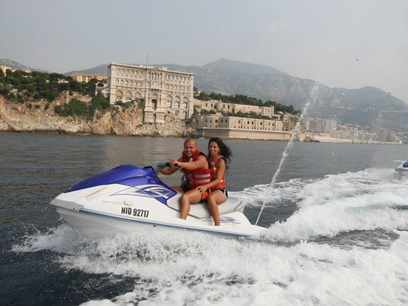 Un couple sur un jetski blanc et bleu navigue près d'une côte avec un grand bâtiment ancien en arrière-plan.