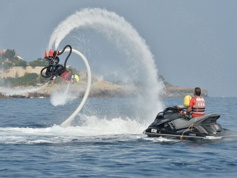 Un homme en gilet de sauvetage pilote un jetski noir tandis qu'une personne utilise un flyboard, projetant de l'eau en arc.