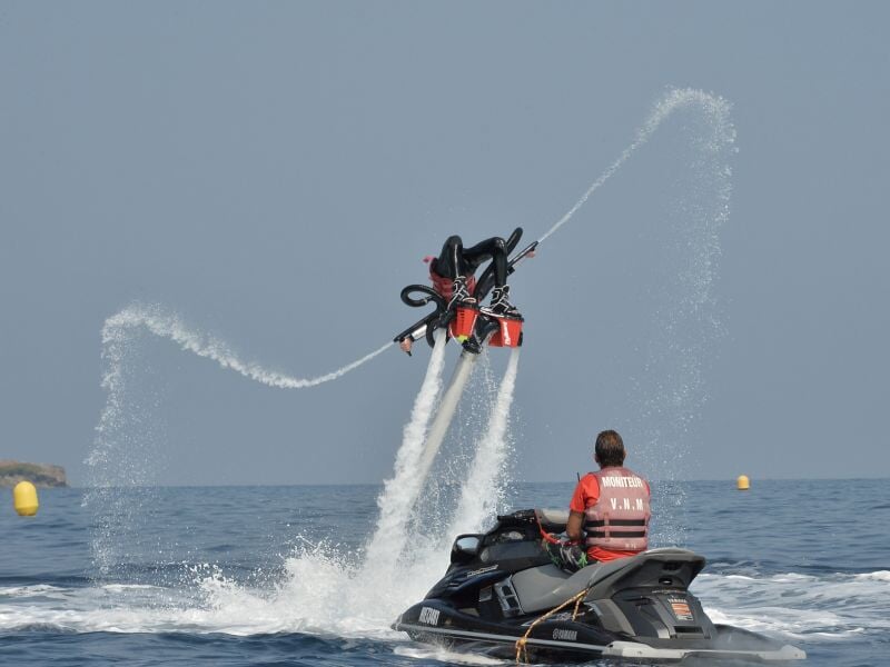 Un homme en gilet de sauvetage sur un jetski noir observe une personne en flyboard qui s'élève au-dessus de l'eau.