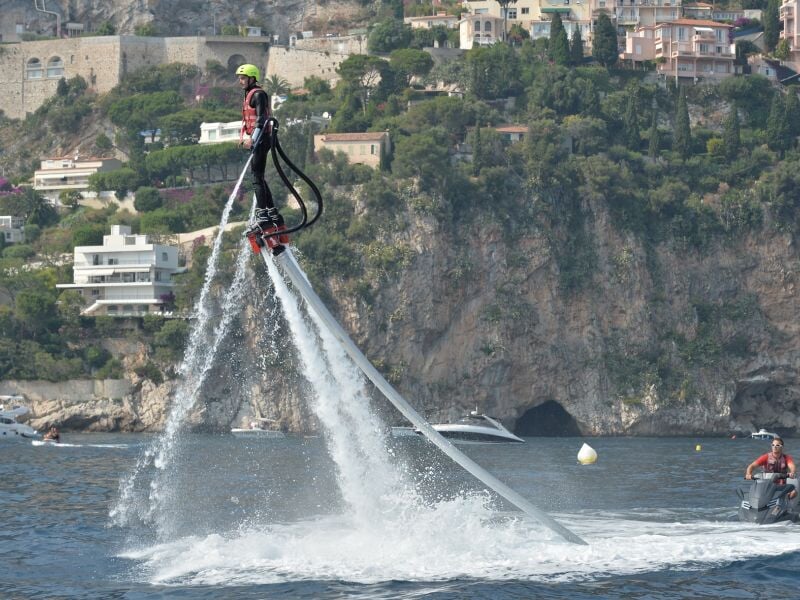 Une personne en combinaison et casque jaune utilise un flyboard pour voler au-dessus de l'eau près d'une côte rocheuse.