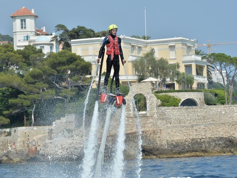 Un homme en gilet de sauvetage sur un jetski noir accompagne une personne en flyboard qui s'élève au-dessus de l'eau calme.