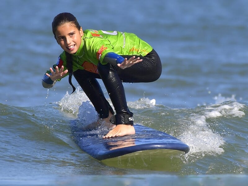 Jeune surfeur en combinaison verte sur une planche bleue, glissant sur une petite vague à La Palmyre.