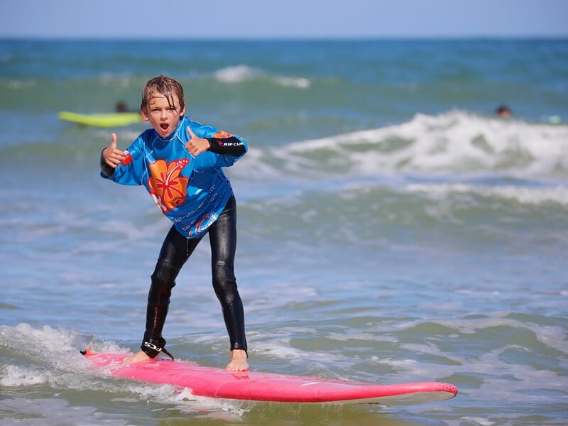 Enfant en combinaison bleue sur une planche rose, surfant une petite vague avec enthousiasme à La Palmyre.