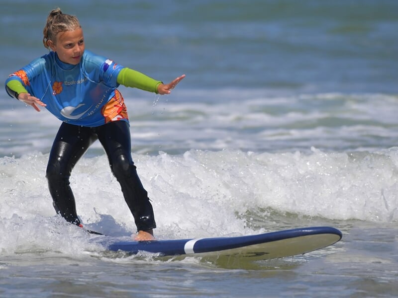 Jeune surfeuse en combinaison bleue et verte, debout sur une planche bleue, surfant une vague douce à La Palmyre.