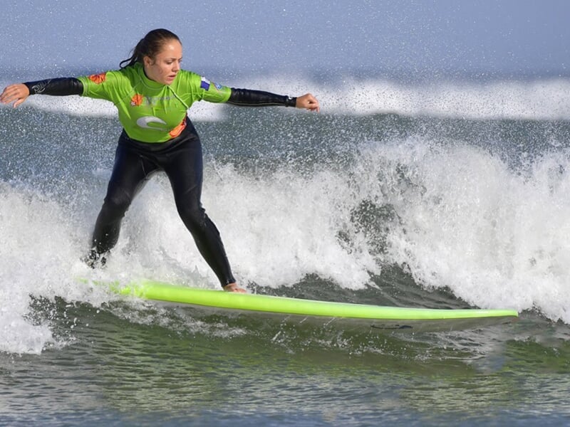Surfeuse en combinaison noire et verte, en équilibre sur une planche verte, surfant une vague à La Palmyre.