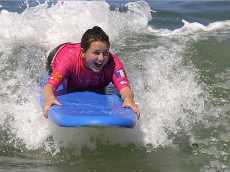 Jeune fille souriante en combinaison rose, allongée sur une planche bleue, surfant une petite vague à La Palmyre.
