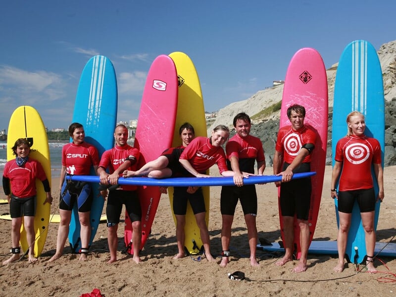 Groupe de jeunes en combinaison rouge posant sur la plage avec planches de surf colorées devant un ciel bleu clair.
