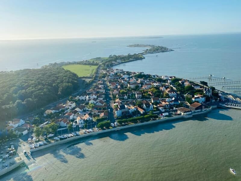 Vue aérienne d'un village côtier avec maisons, forêt dense et mer calme sous un ciel clair bleu.