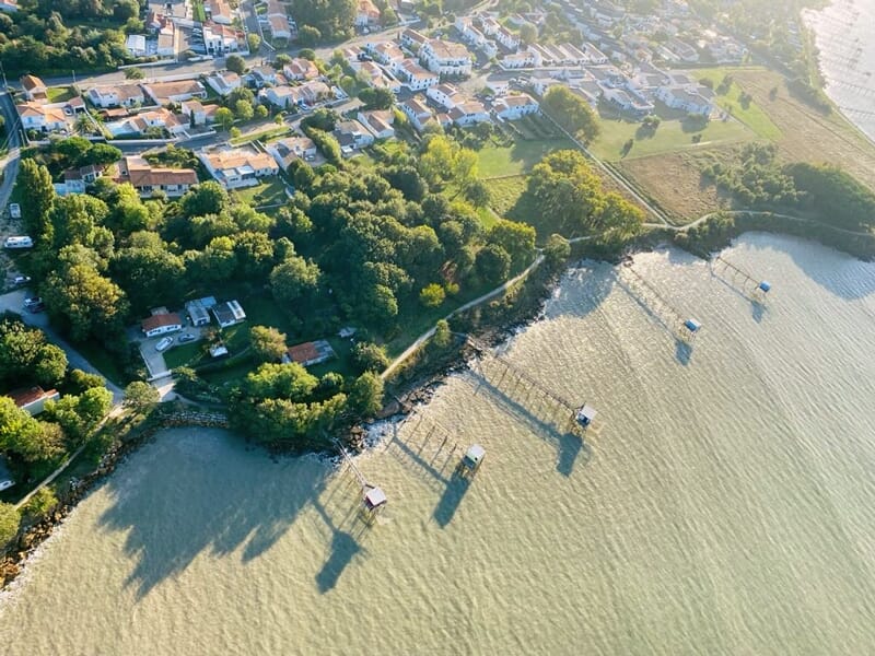 Vue aérienne d'une côte avec des cabanes sur pilotis, arbres verts et eau calme sous un ciel clair.