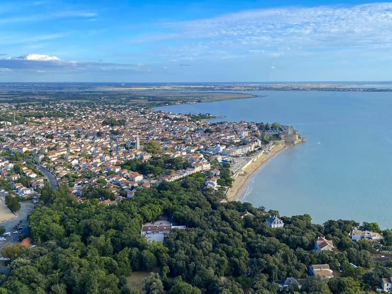 Vue aérienne d'une ville côtière avec maisons, forêt dense et mer sous un ciel bleu clair.