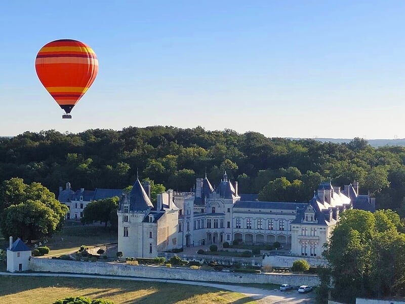 Montgolfière rouge et orange volant au-dessus d'un château entouré d'arbres verts sous un ciel bleu clair.