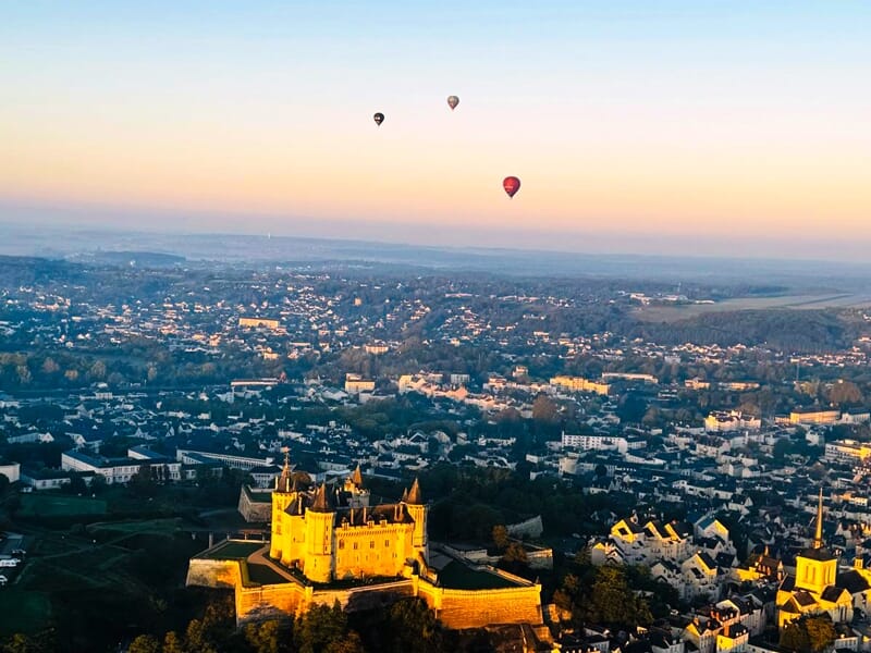 Trois montgolfières flottent au-dessus d'une ville au coucher du soleil avec un château illuminé au premier plan.