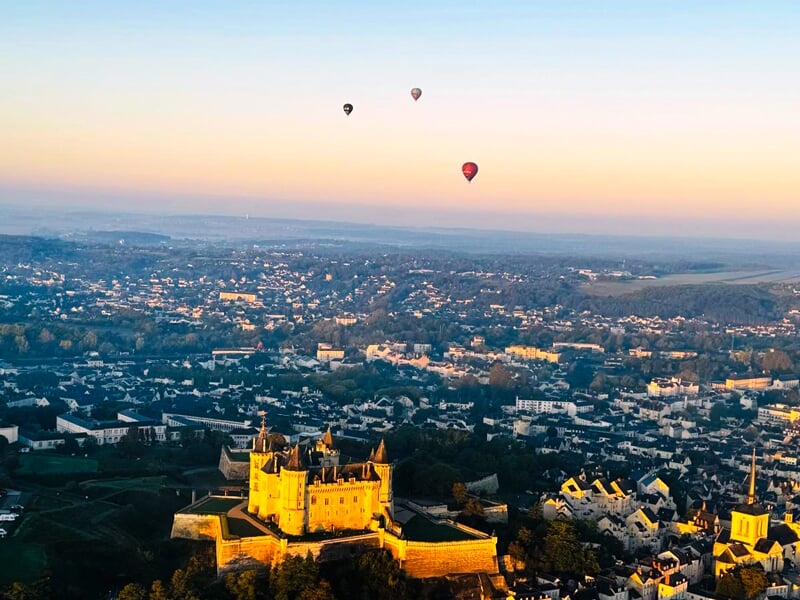 Trois montgolfières flottent au-dessus d'une ville au coucher du soleil avec un château illuminé au premier plan.
