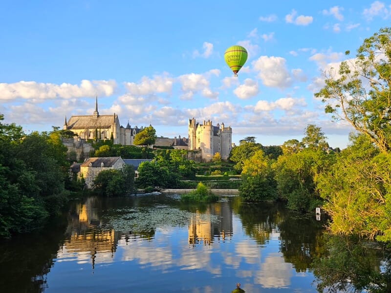 Montgolfière verte survole un château ancien au bord d'une rivière calme reflétant les arbres et le ciel bleu.